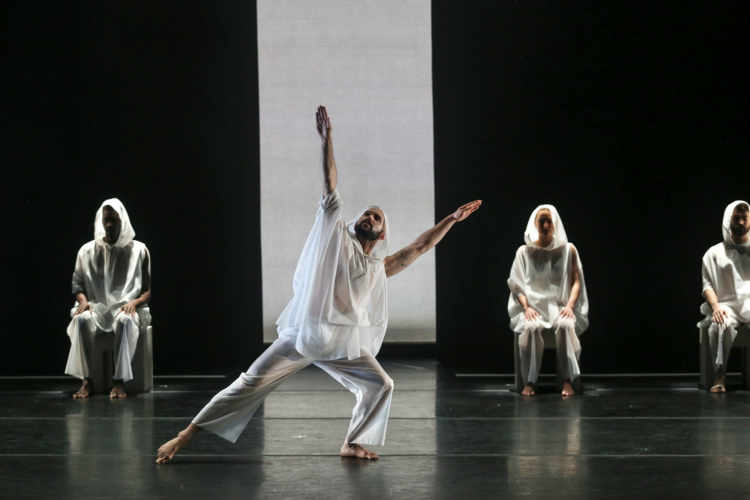 4 figures on a stage - 3 seated in background and 1 standing at center with arms outstretched upward